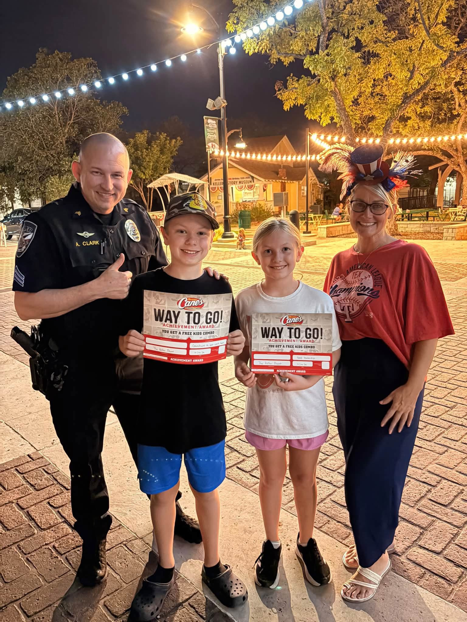 A uniformed police officer, two children holding Raising Cane’s certificates, and an adult wearing a patriotic hat pose on Bates Street at night.